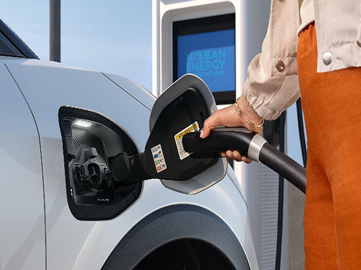 A person plugging a charging cable into the charge port of a white Nissan electric vehicle.